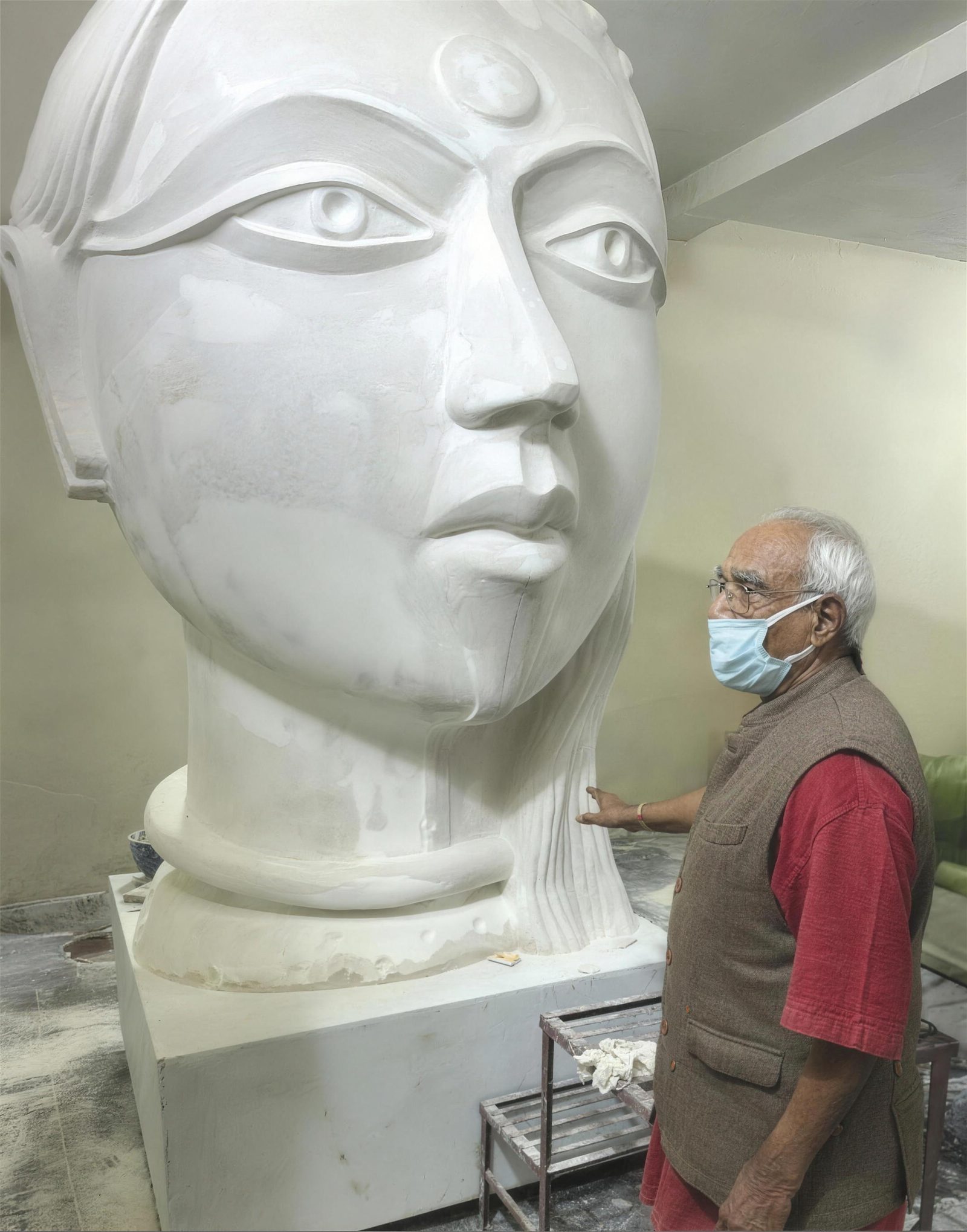 Thota Vaikuntam in a red kurta and mask looking up at a large white marble head under carving in a workshop