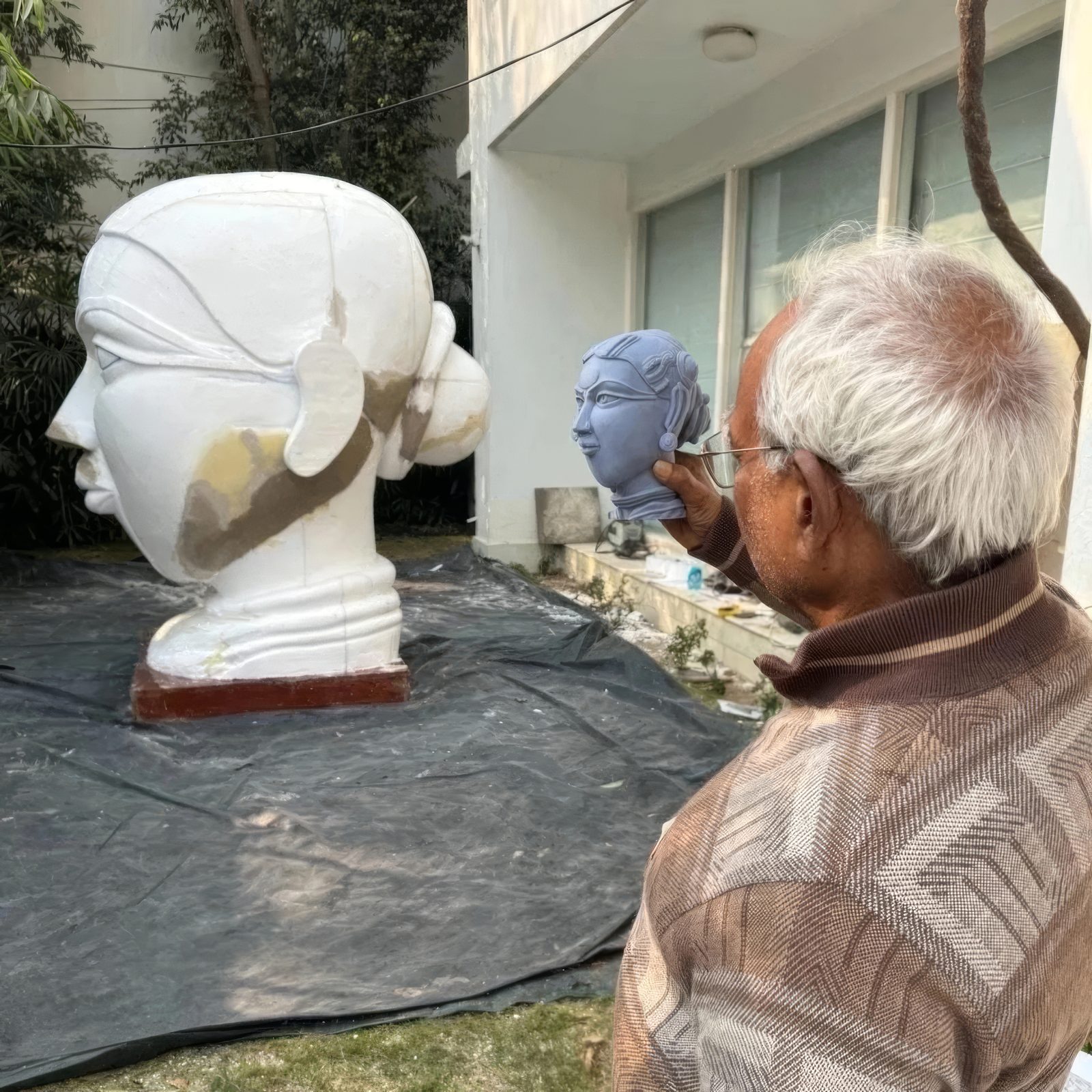 Thota Vaikuntam seen from behind shaping a small head model, with a large white plaster head on a tarpaulin in front of him