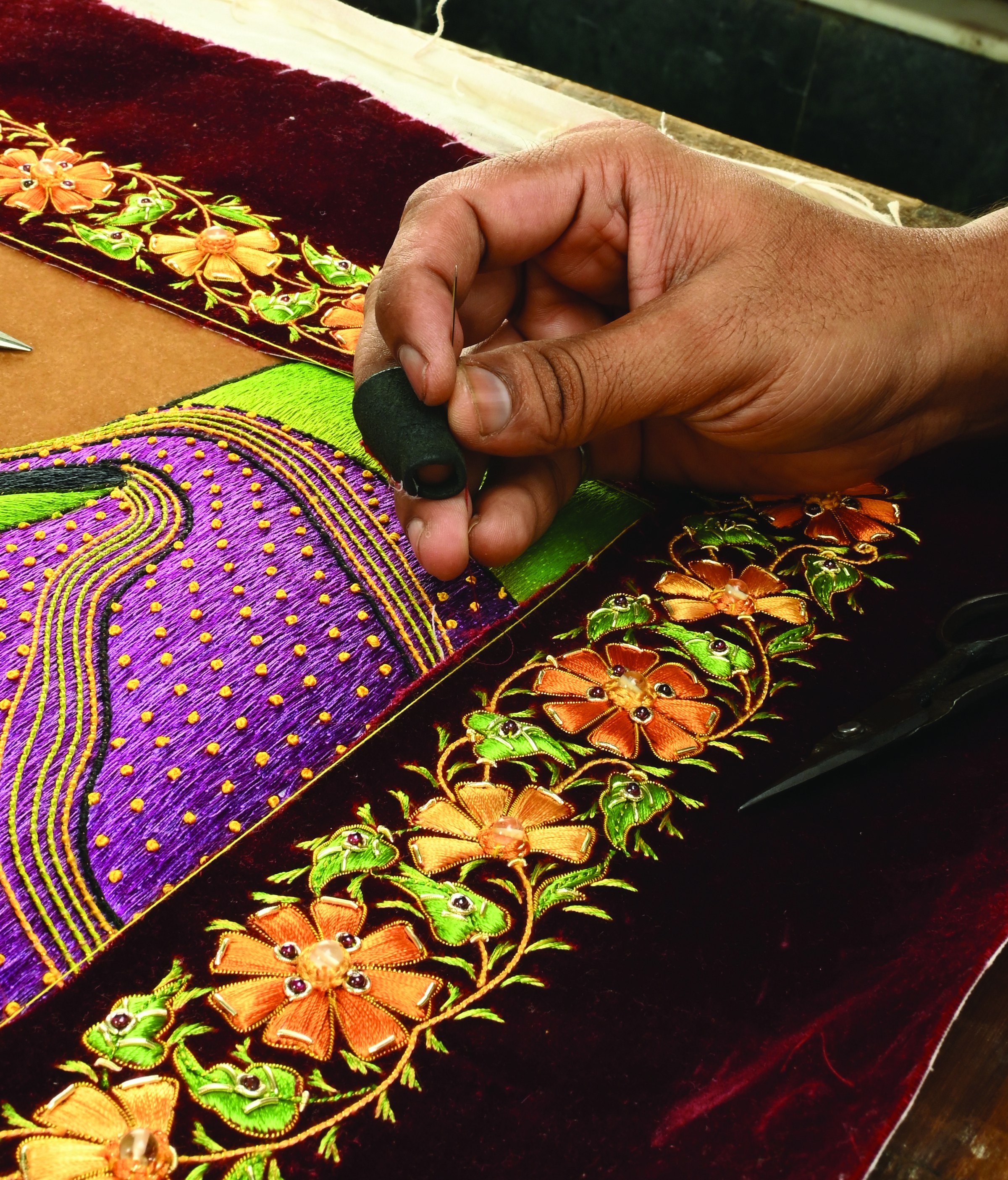 Close-up of a craftsman's hand holding a needle over a maroon velvet zardozi panel with a purple and gold embroidered figure and a floral metal-thread border