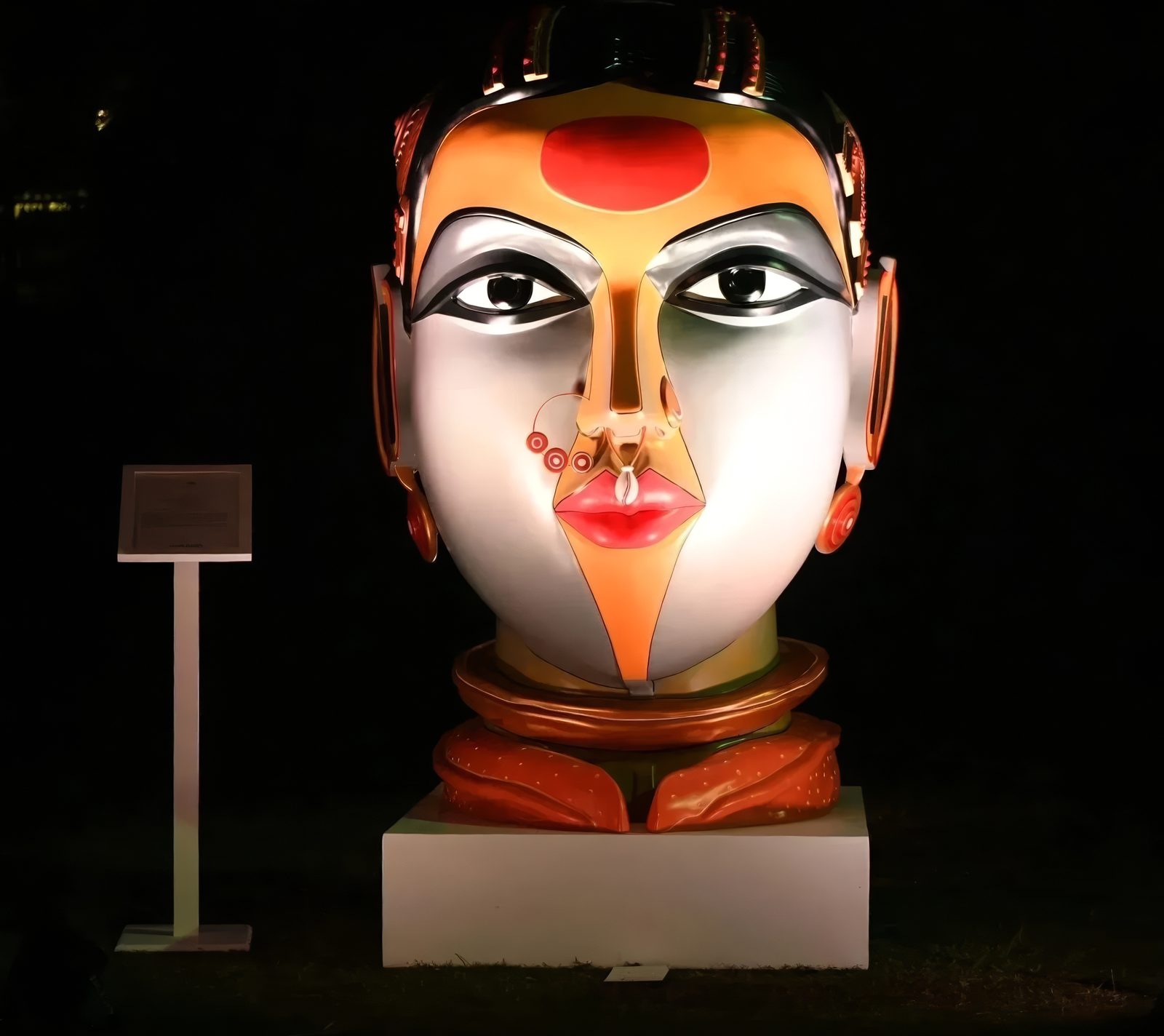 Monumental painted fibreglass head of a Telangana woman by Thota Vaikuntam, silver-grey face with an orange turban and red bindi, lit at night on a white plinth on the lawns of Bikaner House, New Delhi