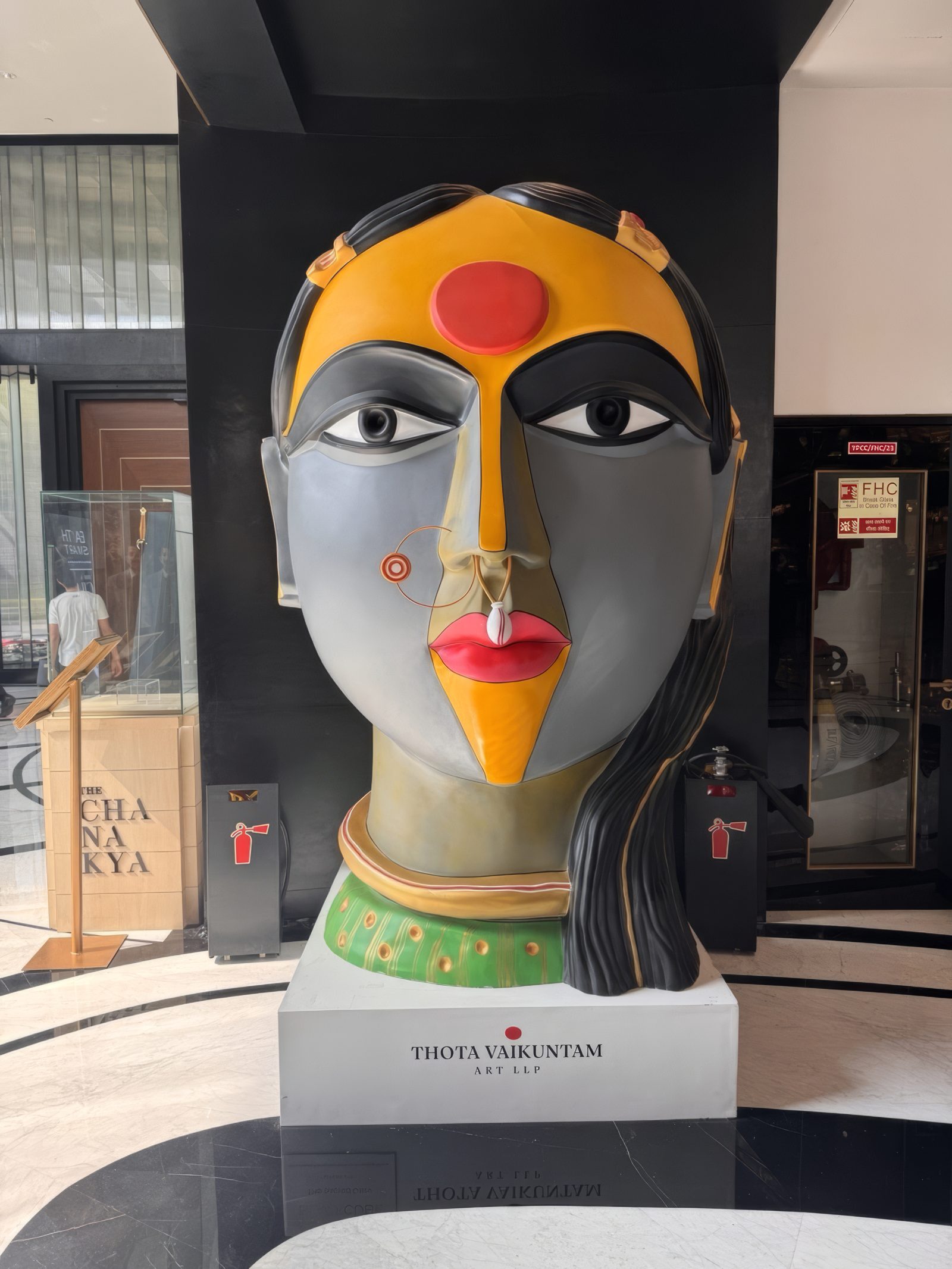 Monumental painted fibreglass head of a Telangana woman by Thota Vaikuntam with a green choker on a white plinth bearing a Thota Vaikuntam plaque, inside The Chanakya, New Delhi