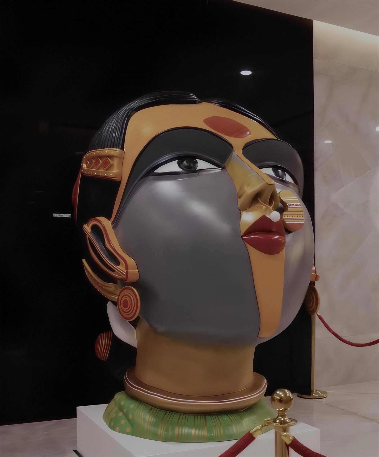 Three-quarter view of a monumental painted fibreglass head of a Telangana woman by Thota Vaikuntam with a gold nose ring, hooped earrings and green choker, installed in a lobby at HITEC City, Hyderabad