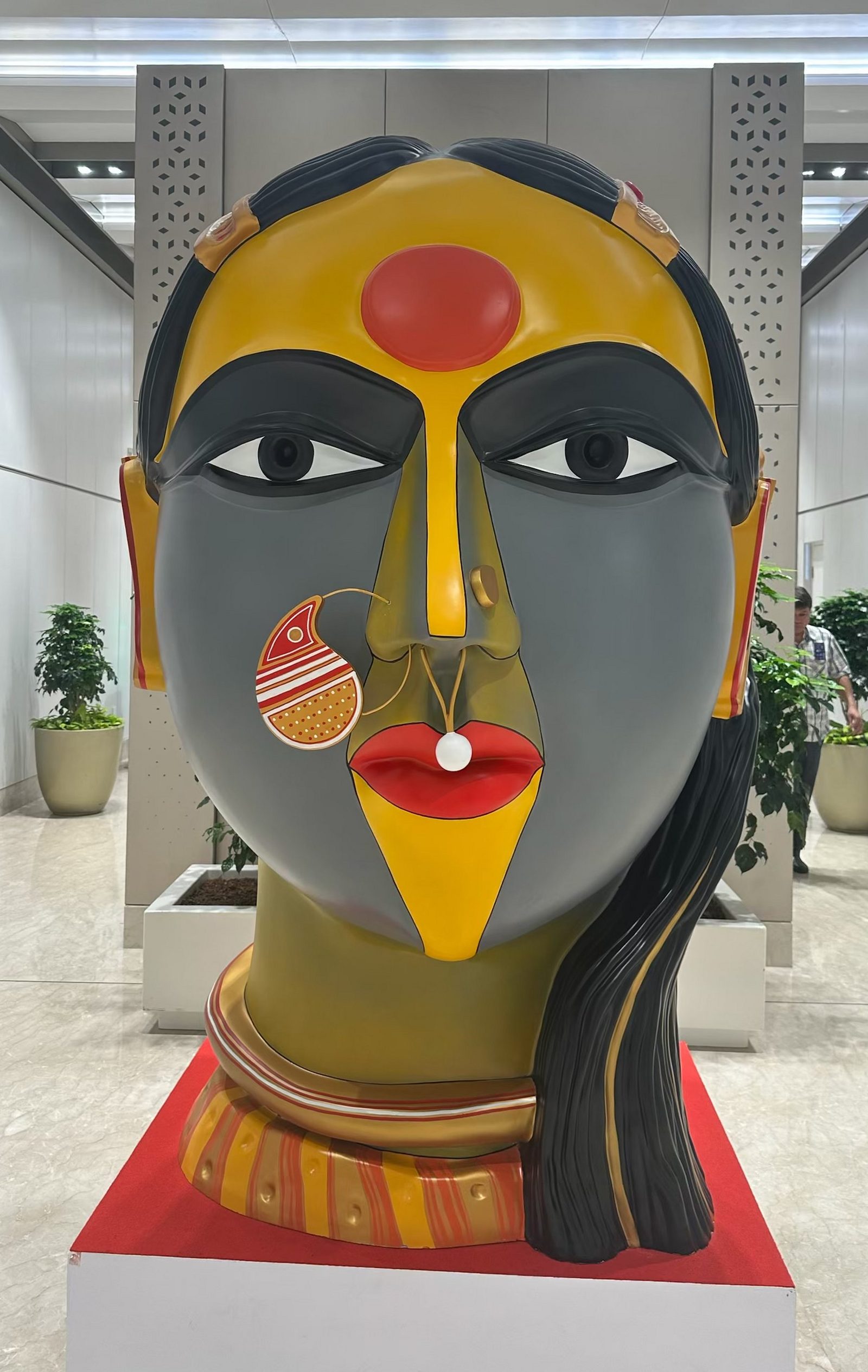 Monumental painted fibreglass head of a Telangana woman by Thota Vaikuntam with a gold nose ring and yellow-and-orange face on a red-topped plinth in the VIP Lounge, Terminal 1, Indira Gandhi International Airport, New Delhi