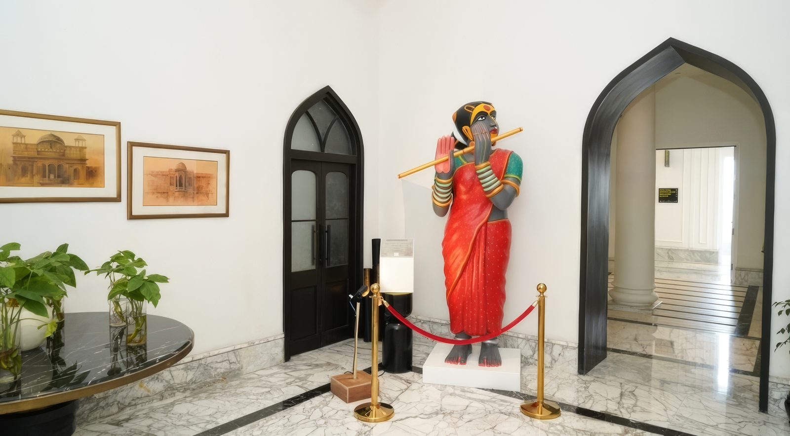 Painted fibreglass sculpture by Thota Vaikuntam of a standing woman in a red sari playing a flute, cordoned by a rope barrier beside an arched doorway at Welcomhotel by ITC Hotels, Amritsar