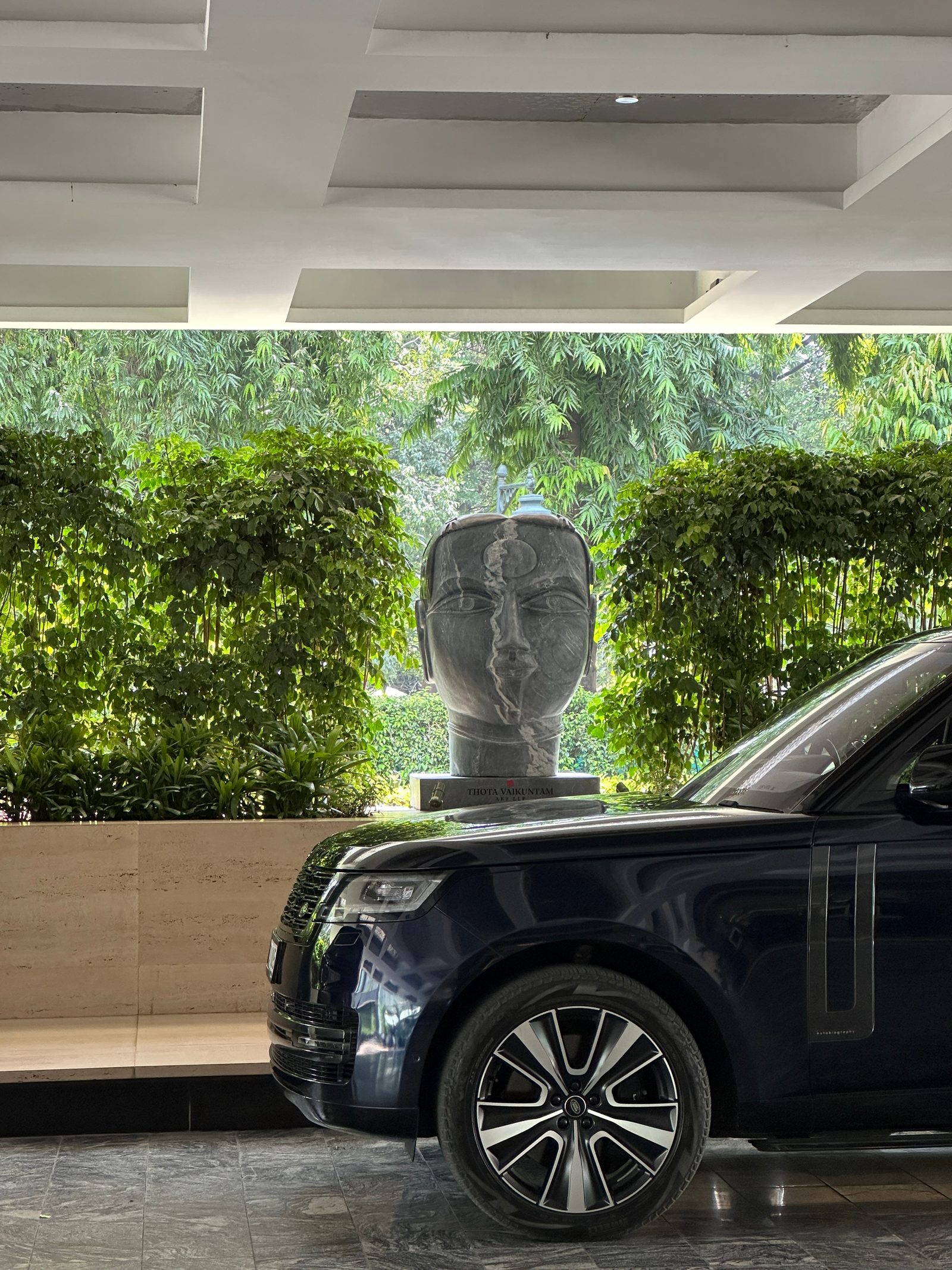 Grey fibreglass head by Thota Vaikuntam set against clipped hedges beside the porte-cochère of Shangri-La Eros, New Delhi, with a black car in the foreground