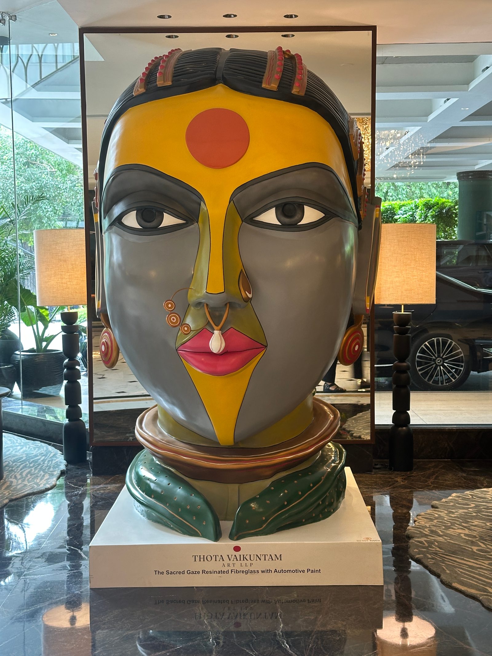 Frontal view of a monumental painted fibreglass head of a Telangana woman by Thota Vaikuntam with a red bindi and nose ring, on a white plinth with a Thota Vaikuntam plaque, in the lobby of Shangri-La Eros, New Delhi